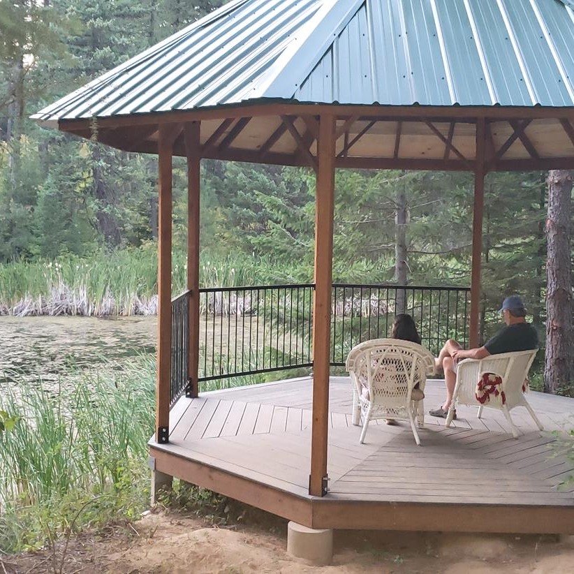 Two people sit on chairs in a gazebo overlooking a pond.